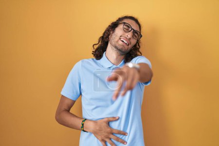 Photo for Young hispanic man standing over yellow background laughing at you, pointing finger to the camera with hand over body, shame expression - Royalty Free Image