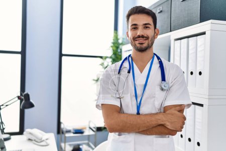 Téléchargez les photos : Young hispanic man wearing doctor uniform standing with arms crossed gesture at clinic - en image libre de droit