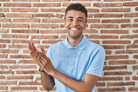 Téléchargez les photos : Brazilian young man standing over brick wall clapping and applauding happy and joyful, smiling proud hands together - en image libre de droit