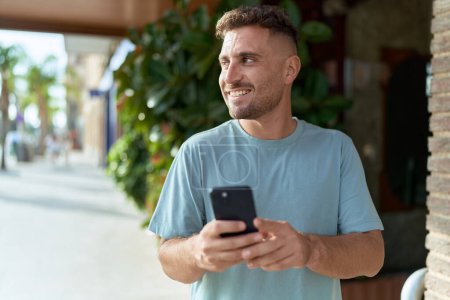 Téléchargez les photos : Jeune homme hispanique souriant confiant en utilisant un smartphone dans la rue - en image libre de droit