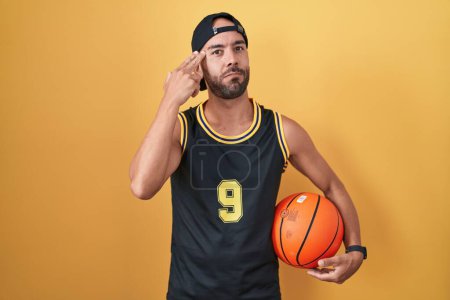 Foto de Hombre calvo de mediana edad sosteniendo la pelota de baloncesto sobre el fondo amarillo disparando y matándose señalando la mano y los dedos a la cabeza como arma, gesto suicida. - Imagen libre de derechos