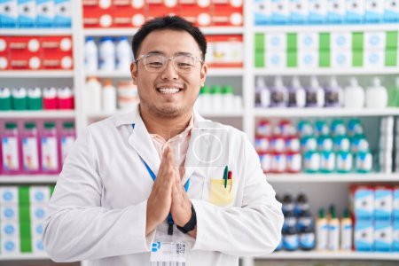 Téléchargez les photos : Jeune homme chinois travaillant à la pharmacie drugstore priant avec les mains ensemble demandant pardon souriant confiant. - en image libre de droit
