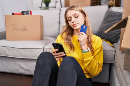 Foto de Mujer rubia joven usando teléfono inteligente y tarjeta de crédito sentado en el piso en el nuevo hogar - Imagen libre de derechos