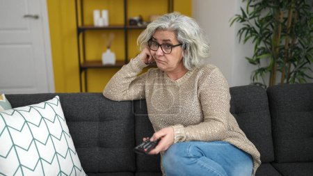 Téléchargez les photos : Femme d'âge moyen avec les cheveux gris regarder la télévision assis sur le canapé avec une expression ennuyeuse à la maison - en image libre de droit
