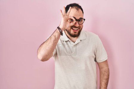 Foto de Hombre hispano de talla grande con barba de pie sobre fondo rosa haciendo buen gesto con la mano sonriente, los ojos mirando a través de los dedos con la cara feliz. - Imagen libre de derechos