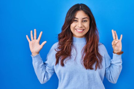 Photo for Hispanic young woman standing over blue background showing and pointing up with fingers number eight while smiling confident and happy. - Royalty Free Image