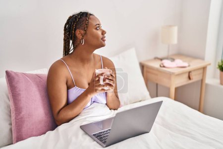 Téléchargez les photos : Femme afro-américaine utilisant un ordinateur portable buvant du café dans la chambre - en image libre de droit