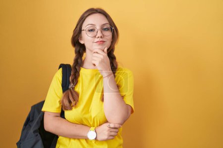 Photo for Young caucasian woman wearing student backpack over yellow background looking confident at the camera smiling with crossed arms and hand raised on chin. thinking positive. - Royalty Free Image