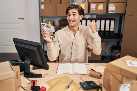 Photo for Young man working at small business ecommerce holding cart pointing thumb up to the side smiling happy with open mouth - Royalty Free Image