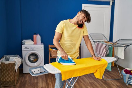 Téléchargez les photos : Young arab man talking on smartphone ironing clothes at laundry room - en image libre de droit