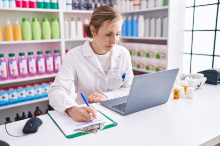 Foto de Joven mujer rubia farmacéutica utilizando la escritura portátil en el documento en la farmacia - Imagen libre de derechos