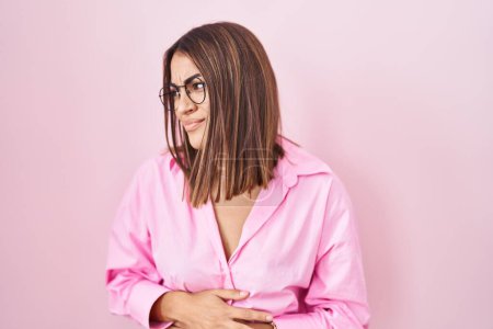 Téléchargez les photos : Jeune femme hispanique portant des lunettes debout sur fond rose avec la main sur le ventre parce que l'indigestion, la maladie douloureuse se sent mal. concept de douleur. - en image libre de droit