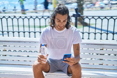 Photo for Young hispanic man using smartphone holding bottle of water at seaside - Royalty Free Image