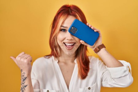 Foto de Mujer joven caucásica cubriendo el ojo con el teléfono inteligente apuntando el pulgar hacia el lado sonriendo feliz con la boca abierta - Imagen libre de derechos
