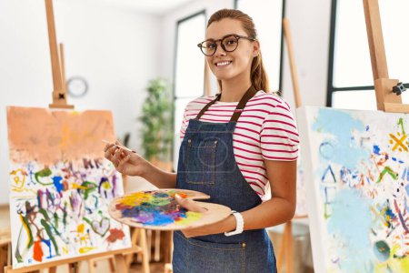 Téléchargez les photos : Jeune femme souriante confiante tenant pinceau et palette au studio d'art - en image libre de droit