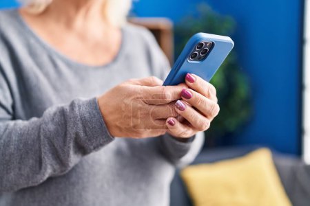 Téléchargez les photos : Middle age blonde woman using smartphone standing at home - en image libre de droit