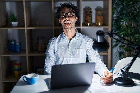 Foto de Hombre hispano trabajando en la oficina por la noche sacando la lengua feliz con expresión divertida. concepto de emoción. - Imagen libre de derechos