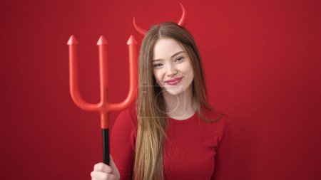 Photo for Young blonde woman wearing devil costume holding trident over isolated red background - Royalty Free Image