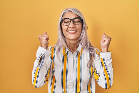 Téléchargez les photos : Femme d'âge moyen avec les cheveux gris debout sur fond jaune portant des lunettes excité pour le succès avec les bras levés et les yeux fermés célébrant la victoire en souriant. concept gagnant. - en image libre de droit