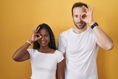 Téléchargez les photos : Couple interracial debout sur fond jaune faisant un geste correct avec la main souriante, les yeux regardant à travers les doigts avec un visage heureux. - en image libre de droit
