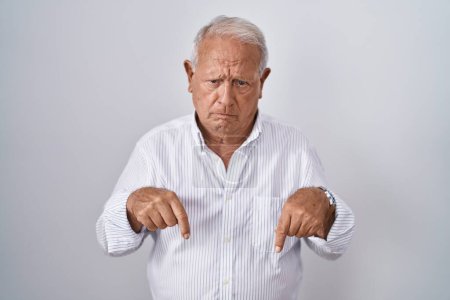 Téléchargez les photos : Homme âgé avec les cheveux gris debout sur fond isolé pointant vers le bas regardant triste et contrarié, indiquant la direction avec les doigts, malheureux et déprimé. - en image libre de droit