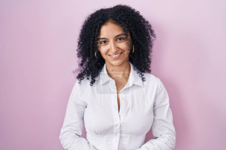 Foto de Hispanic woman with curly hair standing over pink background with hands together and crossed fingers smiling relaxed and cheerful. success and optimistic - Imagen libre de derechos