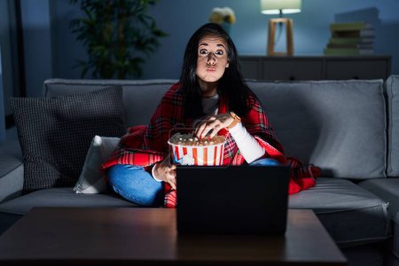 Photo for Hispanic woman eating popcorn watching a movie on the sofa puffing cheeks with funny face. mouth inflated with air, crazy expression. - Royalty Free Image