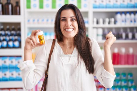 Téléchargez les photos : Jeune femme brune faisant du shopping à la pharmacie pharmacie tenant des pilules criant fier, célébrant la victoire et le succès très excité avec les bras levés - en image libre de droit
