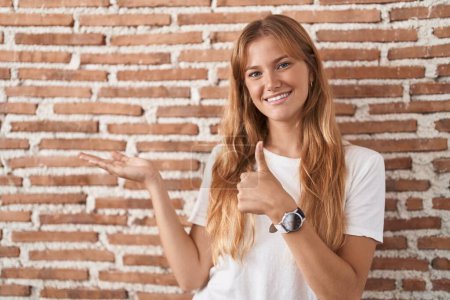 Foto de Joven mujer caucásica de pie sobre la pared de ladrillos que muestra la palma de la mano y haciendo buen gesto con los pulgares hacia arriba, sonriendo feliz y alegre - Imagen libre de derechos