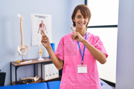 Foto de Brunette woman working at rehabilitation clinic smiling and looking at the camera pointing with two hands and fingers to the side. - Imagen libre de derechos
