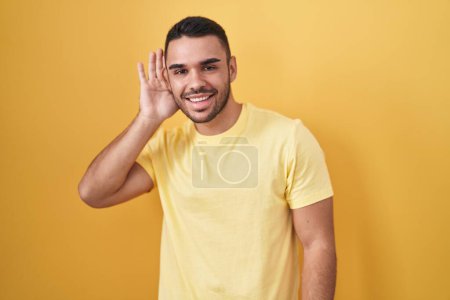 Téléchargez les photos : Jeune homme hispanique debout sur fond jaune souriant avec la main sur l'oreille en écoutant une rumeur ou des commérages. concept de surdité. - en image libre de droit