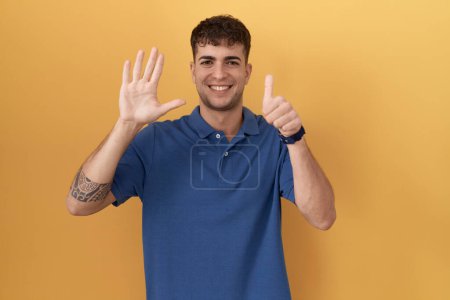Téléchargez les photos : Jeune homme hispanique debout sur fond jaune montrant et pointant vers le haut avec les doigts numéro six tout en souriant confiant et heureux. - en image libre de droit