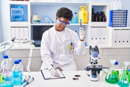 Téléchargez les photos : Femme afro-américaine portant l'uniforme de scientifique écrire sur presse-papiers tenant des tubes à essai au laboratoire - en image libre de droit