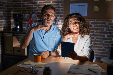 Téléchargez les photos : Couple hispanique moyen-âge à l'aide de touchpad assis sur la table la nuit couper la gorge avec la main comme couteau, menacer d'agression avec une violence furieuse - en image libre de droit