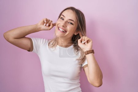 Foto de Blonde caucasian woman standing over pink background smiling pulling ears with fingers, funny gesture. audition problem - Imagen libre de derechos