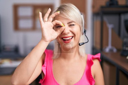Foto de Joven mujer caucásica usando auriculares agente de call center sonriendo feliz haciendo signo de ok con la mano en el ojo mirando a través de los dedos - Imagen libre de derechos