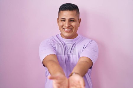 Téléchargez les photos : Young hispanic man standing over pink background smiling with hands palms together receiving or giving gesture. hold and protection - en image libre de droit