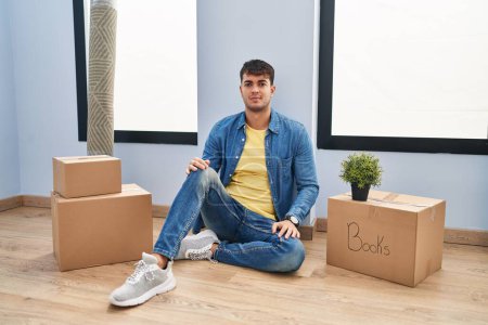 Photo for Young hispanic man sitting on the floor at new home relaxed with serious expression on face. simple and natural looking at the camera. - Royalty Free Image
