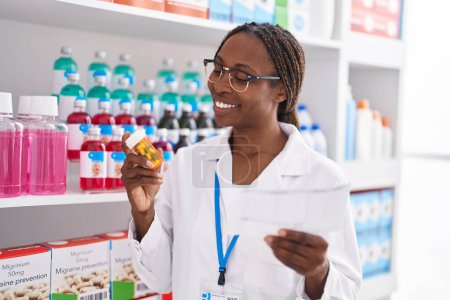 Téléchargez les photos : Femme afro-américaine pharmacien tenant des pilules bouteille lecture prescription à la pharmacie - en image libre de droit