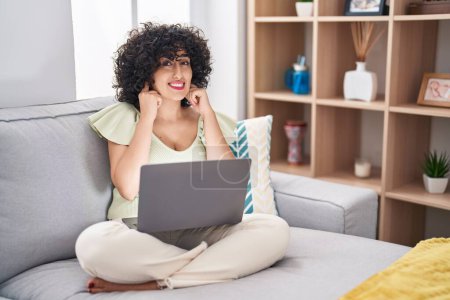 Téléchargez les photos : Jeune femme brune aux cheveux bouclés utilisant un ordinateur portable assis sur le canapé à la maison couvrant les oreilles avec des doigts avec une expression agacée pour le bruit de la musique forte. concept sourd. - en image libre de droit