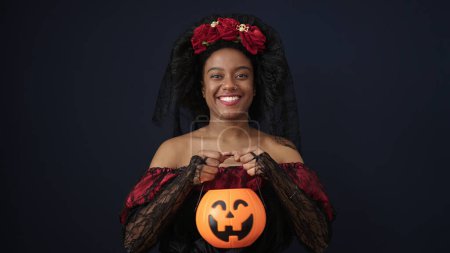 Foto de African american woman wearing katrina costume holding halloween pumpkin basket over isolated black background - Imagen libre de derechos