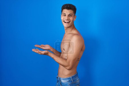 Téléchargez les photos : Jeune homme hispanique debout torse nu sur fond bleu pointant de côté avec les mains ouvertes paumes montrant l'espace de copie, présentant la publicité souriant heureux excité - en image libre de droit