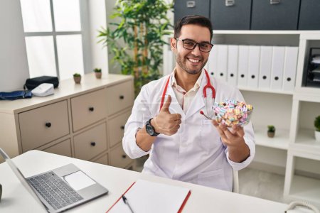 Foto de Joven médico hispano con barba sosteniendo dulces sonriendo feliz y positivo, pulgar hacia arriba haciendo excelente y signo de aprobación - Imagen libre de derechos