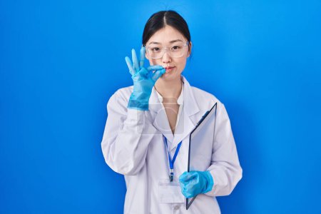 Foto de Chinese young woman working at scientist laboratory mouth and lips shut as zip with fingers. secret and silent, taboo talking - Imagen libre de derechos
