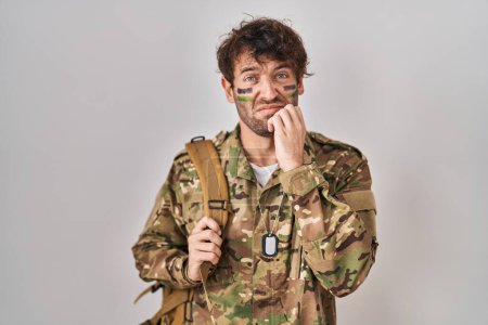 Foto de Joven hispano vistiendo uniforme camuflaje del ejército luciendo estresado y nervioso con las manos en la boca mordiendo uñas. problema de ansiedad. - Imagen libre de derechos