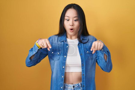 Téléchargez les photos : Jeune femme asiatique debout sur fond jaune pointant vers le bas avec les doigts montrant la publicité, visage surpris et bouche ouverte - en image libre de droit