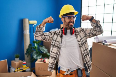 Foto de Joven hombre hispano con barba trabajando en la renovación del hogar mostrando los músculos de los brazos sonriendo orgulloso. concepto de fitness. - Imagen libre de derechos