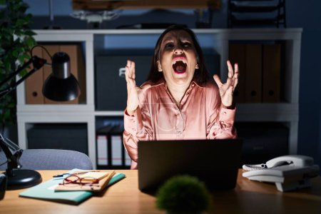 Téléchargez les photos : Femme hispanique du Moyen Age travaillant à l'aide d'un ordinateur portable tard dans la nuit fou et fou crier et crier avec une expression agressive et les bras levés. concept de frustration. - en image libre de droit