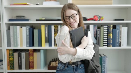Photo for Young blonde woman student standing hugging book at university classroom - Royalty Free Image