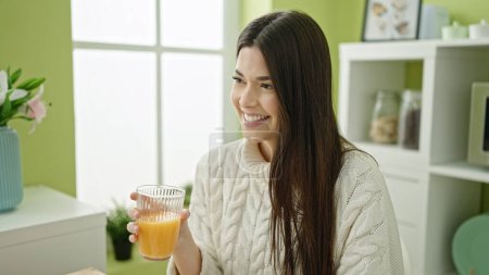 Téléchargez les photos : Jeune belle hispanique femme boire du jus d'orange assis sur la table à la maison - en image libre de droit